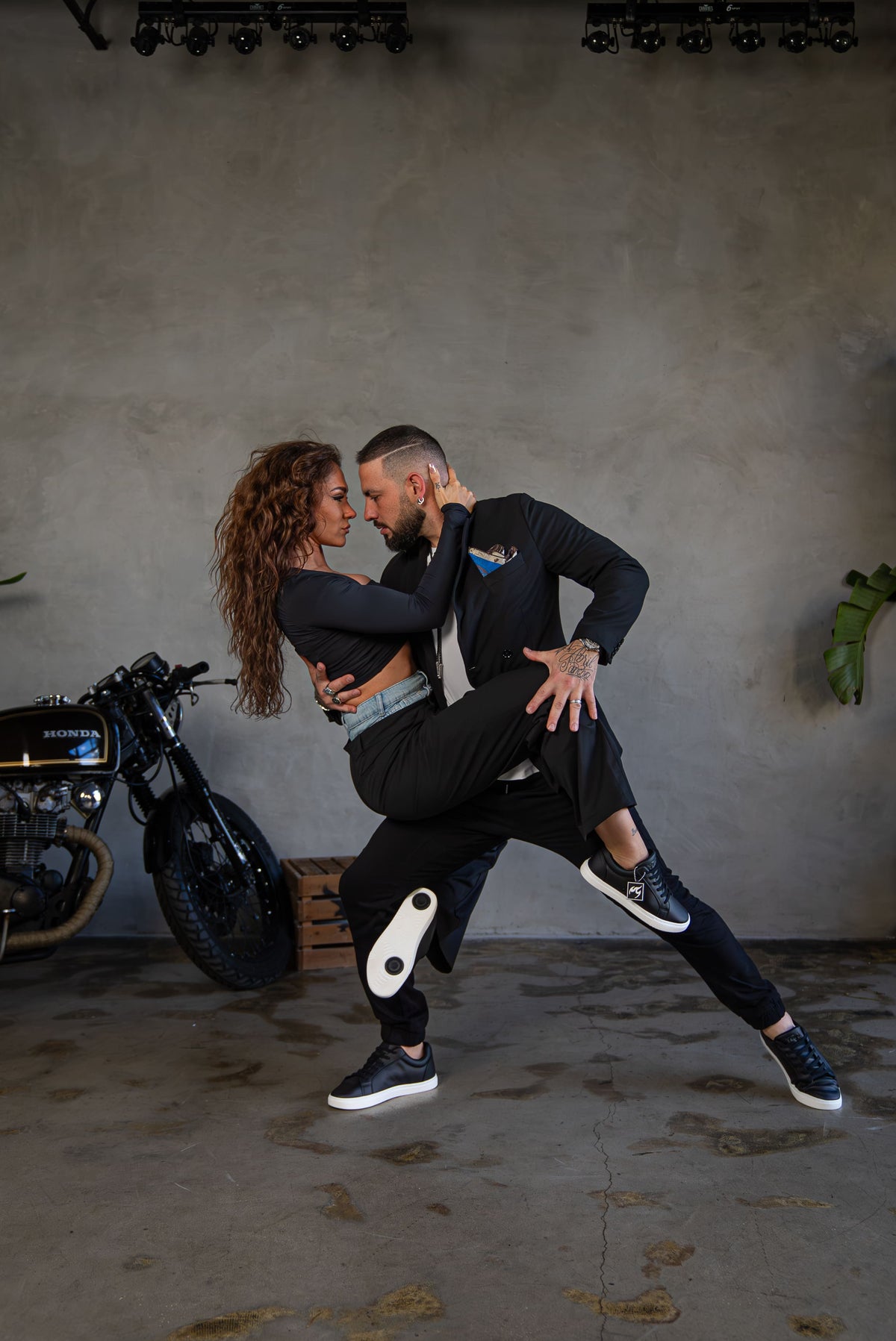 A man and a woman dance closely in a dim indoor setting with a concrete wall, industrial ceiling lights, and a motorcycle at the left edge. The man wears a black long-sleeve top and black pants with white sneakers, while the woman wears a black crop top and black pants. Fuego Lowtop Black dance salsa bachata.